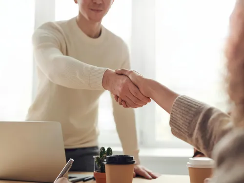 a man standing shakes the hand of another person with a desk, coffees and a laptop in betweenHappy to 